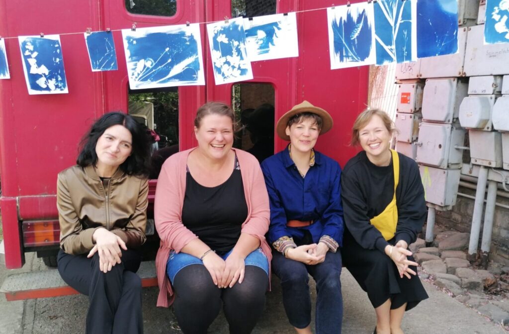 Vier Frauen sitzen lachend vor einem roten Bahnwaggon, über ihnen hängt eine Leine mit blau-weißen Cyanotypie-Drucken mit Pflanzenmotiven. Four women sit laughing in front of a red railway carriage, with a line of blue-and-white cyanotype prints featuring plant motifs hanging above them.