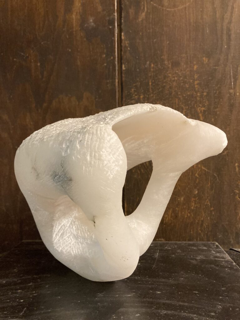 Abstrakte Skulptur aus durchscheinend weißem Alabaster mit organisch geschwungener Form und ausgehöhlter Öffnung, fotografiert vor dunklem Holzuntergrund.